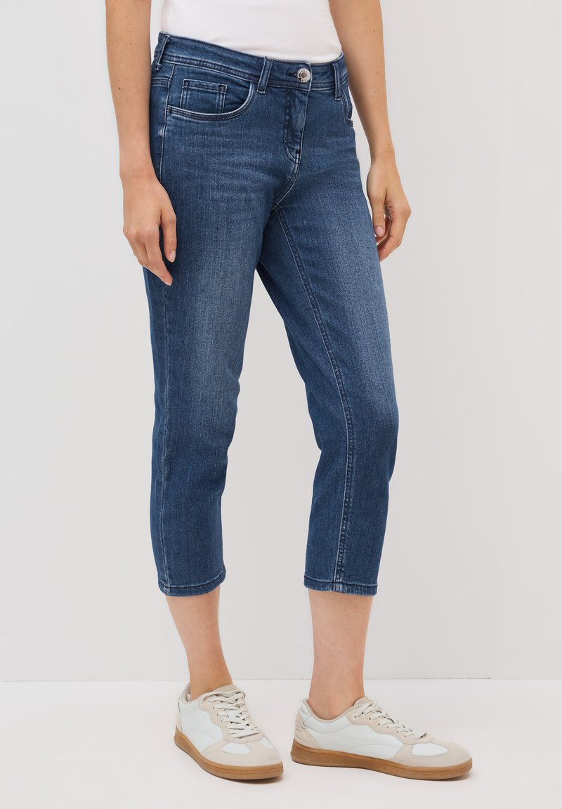 Pantalons capri en denim bleu avec une coupe ajustée, dotés de poches avant, d'une fermeture à bouton classique et d'ourlets retroussés. Portés avec des baskets blanches.