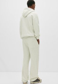 Person wearing light gray hoodie and matching sweatpants with hands in pockets, standing against plain white background, facing away.