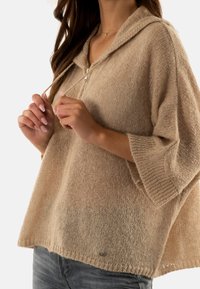 Pull à capuche beige avec coupe décontractée, tricot texturé, fermeture à cordon, et manches courtes, associé à un jean bleu clair.