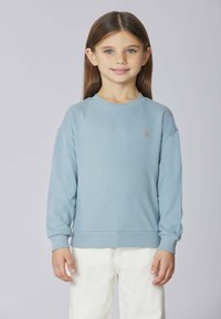 Ragazzina con lunghi capelli castani, che indossa una felpa azzurra chiara e pantaloni bianchi, in piedi contro uno sfondo grigio chiaro semplice.