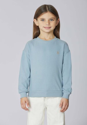 Ragazzina con lunghi capelli castani, che indossa una felpa azzurra chiara e pantaloni bianchi, in piedi contro uno sfondo grigio chiaro semplice.