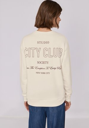 Felpa color crema con grande testo marrone sul retro: "CITY CLUB" e "NEW YORK CITY", con ulteriore testo sotto. Vestibilità rilassata e maniche lunghe.