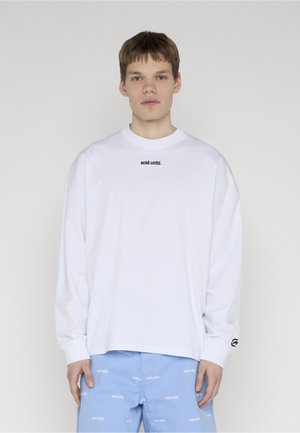 Tânăr privindu-se înainte, purtând o cămașă albă cu mâneci lungi inscripționată cu textul „ecko unltd.” și pantaloni albastru deschis cu un model repetat de text.