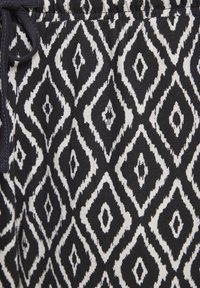 Schwarz-weißer gemusterter Stoff mit einem Diamantdesign und gezackten Kanten. Das Material wirkt strukturiert und hat einen elastischen Bund.