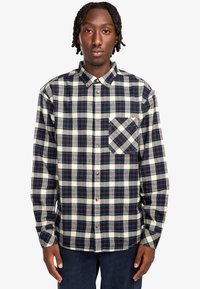 Camisa de franela de cuadros en navy, crema y burdeos, con cuello abotonado, botones y un bolsillo de parche con patrón de cuadros en diagonal.