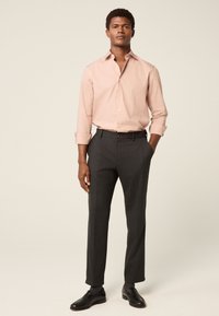 Svijetloružičasta košulja na kopčanje s podvrnutim rukavima u kombinaciji s hlačama boje ugljena i crnim mokasinama, naglašavajući formalni, krojeni izgled.