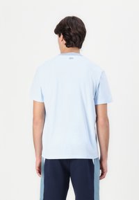 Tricou din bumbac de culoare albastru deschis, cu guler rotund și mâneci scurte. Prezintă un logo subtil deasupra gulerului. Textură netedă, croială casual.