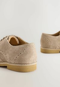 Brogues en suède beige avec perforations décoratives, texture lisse et semelle en caoutchouc brun clair. Dotés de lacets et d'un bout rond.