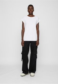 Weißes T-Shirt mit kurzen Ärmeln, kombiniert mit schwarzen Cargo-Hosen mit seitlichen Taschen und Reißverschlüssen, getragen mit hellfarbenen Schuhen.