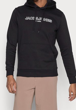 Schwarzer Kapuzenpullover mit sichtbarem weißen "JACK & JONES"-Logo und Fronttasche, getragen von einer Person mit beigen Hosen vor einem einfarbigen Hintergrund.