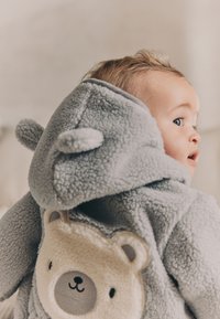 Pelēkā flīsa kapuce mēteli ar lāča dizainu mugurā, aprīkota ar noapaļotām ausīm, maigu tekstūru un krēmkrāsas vēderu.