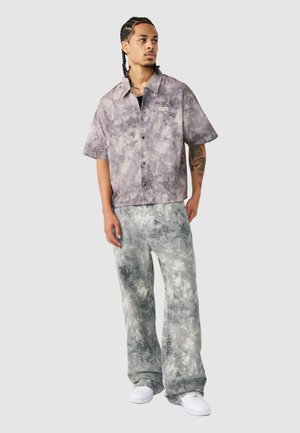 Man met gevlochten haar, die een grijs tie-dye shirt met korte mouwen en bijpassende wijde broek draagt, staand en naar de zijkant kijkend.