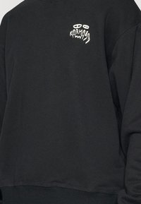 Schwarzer Sweatshirt aus Baumwollmischung mit einem weißen Grafiklogo auf der Brust. Er hat eine lockere Passform und gerippte Bündchen sowie einen gerippten Saum.