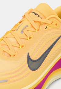 Αθλητικό παπούτσι Nike με φωτεινό κίτρινο επάνω μέρος από δίχτυ με υφασμάτινες λεπτομέρειες, στρωμένη σόλα σε μωβ και κίτρινο, και εμφανές μαύρο λογότυπο swoosh.