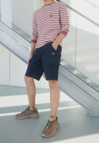 Muž oblečený v červeném a bílém pruhovaném tričku s dlouhým rukávem, tmavě modrých šortkách s kapsami a béžových sportovních botách, stojící u kovových schodů.