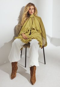 Olivza zaņķota tunika, baltas platās bikses un brūni zamšādas botas ar norādītu purnu. Melns piekaramā rotaslieka akcentē apģērbu.