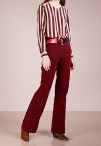Blouse à manches longues avec des rayures verticales dans des nuances de bordeaux, crème et marron. Associée à un pantalon flare en velours côtelé bordeaux et une ceinture rouge.
