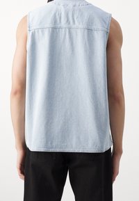 Světle modrá denimová košile bez rukávů s zadním dílem, rovný lem a neupravené okraje. Kombinováno s černými kalhotami a ukazující detaily na rameni a pažích.