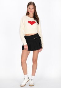Crème cropped sweater met een rood hartontwerp, gecombineerd met een zwarte geplooide mini-jurk met een zijriem en gespsluiting. Witte sneakers en sokken.