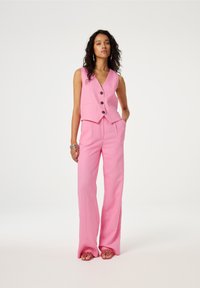 Roze linnen vest met een V-hals en drie knopen, gecombineerd met wijde roze broeken. Sandalen met open tenen en bloemenmotieven.