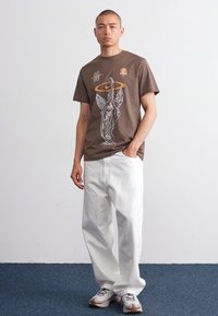 Brun bomull T-skjorte med grafisk trykk som viser en engel og sirkeldesign. Kombinert med løstsittende hvite denimbukser og sneakers.