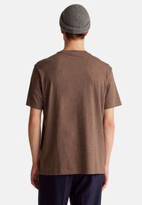 T-shirt marrone a maniche corte con colletto girocollo, realizzato in un tessuto morbido. Indossato con pantaloni scuri, visto da dietro, con un berretto grigio.