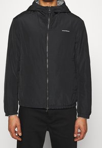 Svart vindjacka med dragkedja fram, elastisk nederkant och ett mönstrat innerfoder i huven. Har sidosfickor och en snygg design.