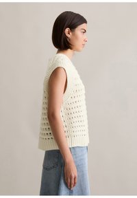 Haken mouwloos sweater in crème met open gaten, geribde halslijn en zoom, gecombineerd met een spijkerrok in denimblauw, zijaanzicht weergegeven.