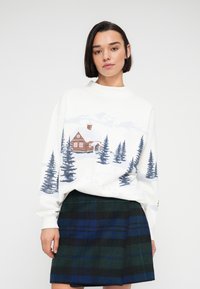 Γυναίκα που φοράει λευκό πουλόβερ με σχέδιο χιονισμένης καλύβας και πεύκων, συνδυασμένο με καρό φούστα σε μπλε και πράσινους τόνους, στέκεται μπροστά σε απλό φόντο.