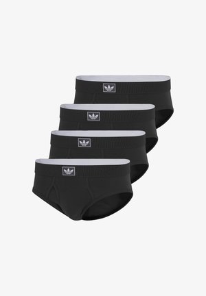 Umięśniony mężczyzna stoi boso, ubrany w czarne markowe slipy, patrząc w bok na tle jednolitego białego tła.