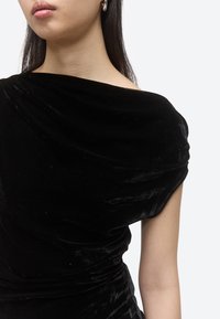 Nő fekete bársonyos, vállat szabadon hagyó felsőt visel, rajta redőzött anyag és ezüst karika fülbevalók, egyszínű háttér előtt.