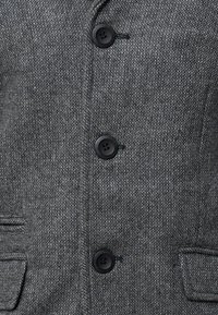 Manteau en laine grise avec tissu texturé, cinq boutons noirs et deux poches latérales. Coupe ajustée avec un col classique.