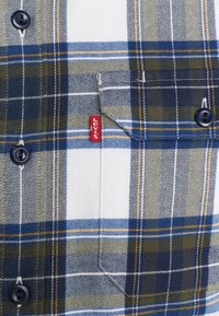 Modrá, zelená a bílá kostkovaná košile s tmavými knoflíky, má přední kapsu a červenou nášivku na oblasti hrudníku. Bavlněná tkanina, ležérní design.