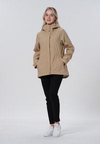Beige wasserdichte Jacke mit Kapuze, elastischem Bund, zwei seitlichen Taschen und Druckknopfverschlüssen. Kombiniert mit schwarzen Hosen und weißen Turnschuhen.