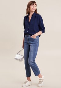 Marineblauw overhemd met opgerolde mouwen, lichtblauwe straight-leg jeans, zilveren crossbody tas en witte sneakers met grijze strepen.