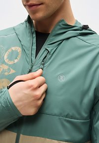 Άνδρας που κλείνει ένα πράσινο και μπεζ μπουφάν με κουκούλα, με αφηρημένα σχέδια και λογότυπο στο στήθος, φοράει μαύρο πουκάμισο από κάτω.