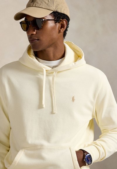 Giovane uomo con cappellino beige, occhiali da sole neri, felpa giallo pallido con logo e orologio da polso con quadrante blu, che guarda di lato.