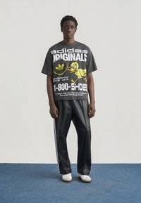 Άνδρας που στέκεται φορώντας μαύρο T-shirt Adidas Originals με κίτρινο γραφικό πάπιας και μαύρο παντελόνι με λευκές ρίγες, πάνω σε μπλε χαλί.