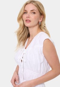 Rochie albă din bumbac cu mâneci scurte, cu un corset plisat și fundiță la decolteu. Detalii brodate adaugă textură și interes vizual.