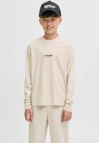Junge trägt ein beiges Langarmshirt mit dem Text "JJ Studios", passende beige Hosen und eine schwarze Kappe mit dem Text "Originals", lächelnd vor einem einfarbigen Hintergrund.