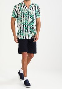 Kurzärmliges Hemd in Grün und Weiß mit Palm- und Streifenmustern; kombiniert mit dunklen Shorts und marineblauen Sneakers. Glatte Stofftextur.