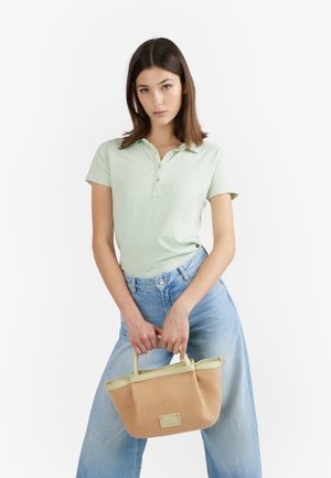 Giovane donna con polo verde chiaro e jeans blu che tiene una borsa intrecciata beige con entrambe le mani, su sfondo bianco.