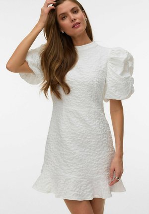 Femme aux longs cheveux bruns portant une robe blanche texturée avec des manches courtes bouffantes et un ourlet à volants, touchant ses cheveux.