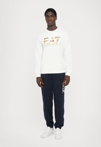 Sweatshirt branco com o logótipo dourado "EA7", combinado com calças de treino azul-marinho com detalhes brancos e ténis brancos. O modelo está de pé contra um fundo simples.