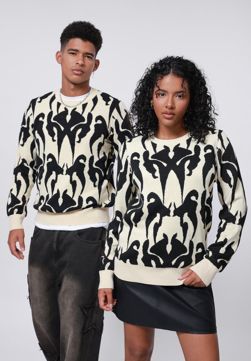 Strickpullover in Creme mit schwarzen abstrakten Mustern, ausgestattet mit gerippten Bündchen und Saum. Modell kombiniert mit einem schwarzen Kunstlederrock und Jeans.
