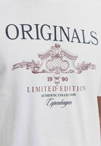 Biela tričko s čiernym a hnedým vintage štýlovým textom a ornamentom, ktorý znie "ORIGINALS 1990 LIMITED EDITION AUTHENTIC COLLECTION Kodaň."
