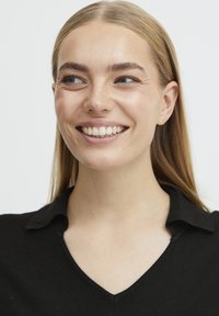 Fekete kötött felső V-kivágással és gallérral. A modellen egyenes, világosbarna haj van, és semleges arckifejezése mellett láthatóak a fogai.