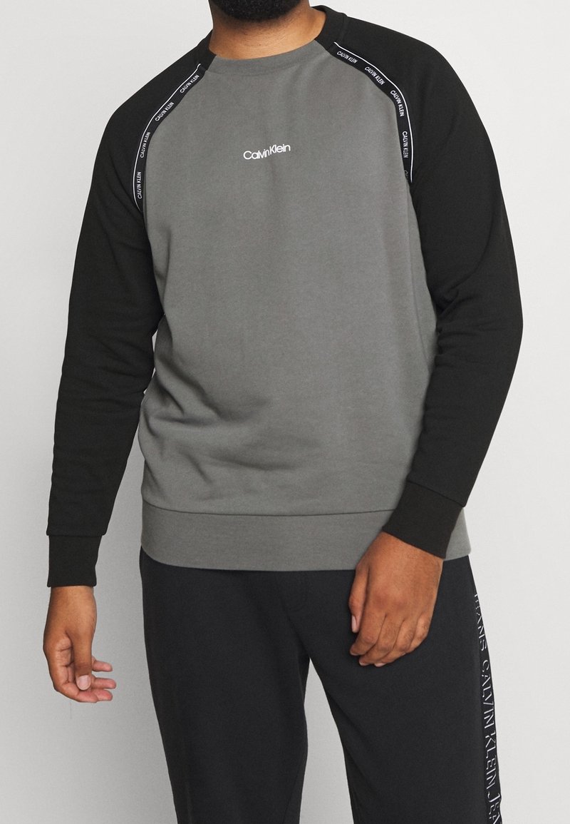 Homme portant un sweat-shirt Calvin Klein gris et noir avec le logo sur la poitrine et un ruban de marque sur les épaules, assorti à un pantalon noir.