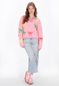 Rosa, flauschiger Pullover mit mehrfarbigen Schleifenverzierungen, V-Ausschnitt, kombiniert mit einem hellen Jeansrock mit seitlichem Schlitz und rosa Turnschuhen.