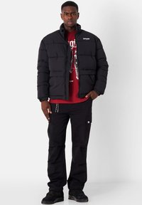Giacca a vento nera con chiusura a zip e cuciture orizzontali. Indossata sopra una t-shirt grafica rossa e abbinata a pantaloni cargo neri e scarpe.